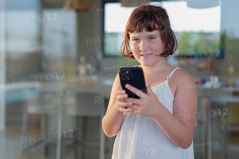 Young girl enjoying a sunny day outdoors while engaging with her smartphone. She is relaxed, standing in a sunlit area, dressed in light summery clothing, capturing a tech-savvy lifestyle