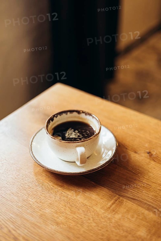 A ceramic coffee cup filled with rich black coffee, topped with delicate latte art foam, rests on a wooden table, creating a warm and inviting atmosphere in a cafe