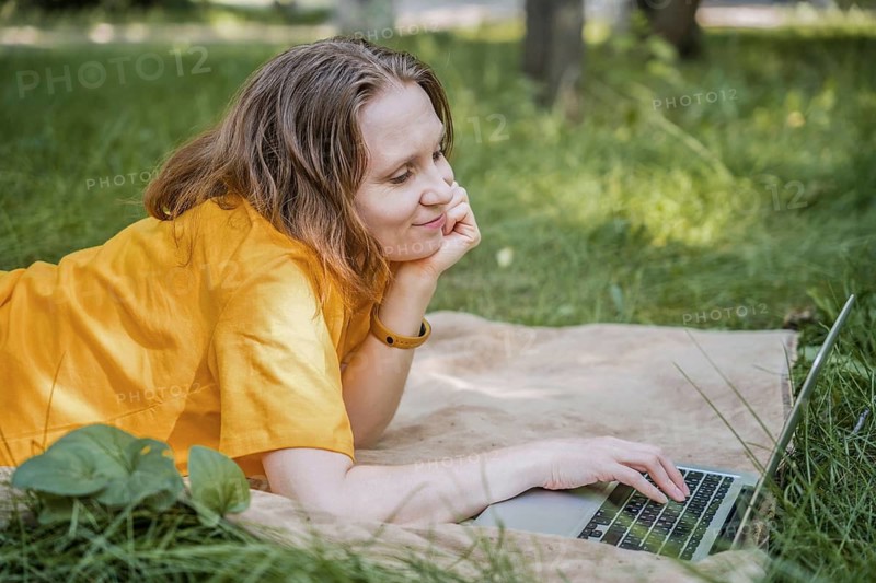 An happy woman in a yellow shirt lies in the grass at a park, working on a laptop. She is smiling, enjoying a remote work setting in a natural, relaxed environment