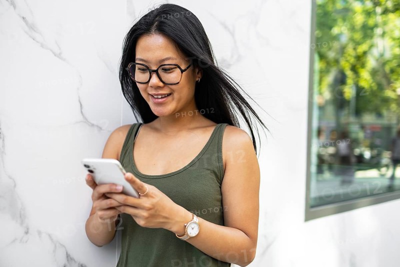 A young Chinese woman stands outdoors in a summer city setting, smiling as she uses her smartphone. She is casually dressed, enjoying a sunny day in the urban environment