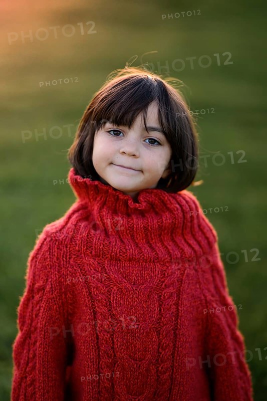 A young girl with a joyful expression, wearing a red cable-knit sweater, stands outdoors with a softly blurred autumn sunset in the background. She epitomizes the warmth and charm of a crisp autumn evening