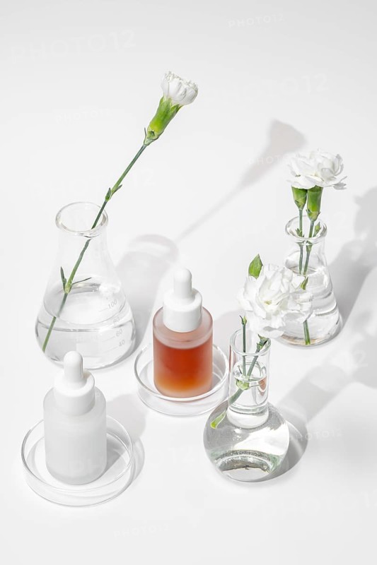 From above serene arrangement of modern skincare products alongside elegant flowers in various scientific glassware, reflecting purity and innovation in skincare development against white background