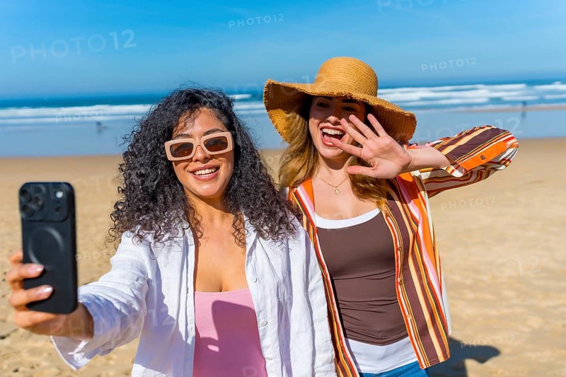 Two cheerful young women taking a selfie on a sunny sandy beach, laughing and posing by the ocean waves while enjoying a carefree summer vacation together