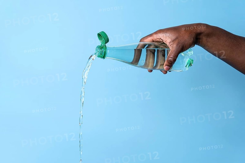 A close-up of a hand pouring water from a clear plastic bottle against a light blue background. The image captures the fluid motion and transparency of the flowing water