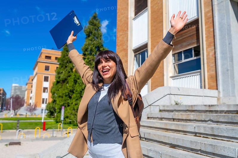 Happy young woman raising arms in front of a university building, celebrating a success or achievement, representing education, winning, and new beginnings