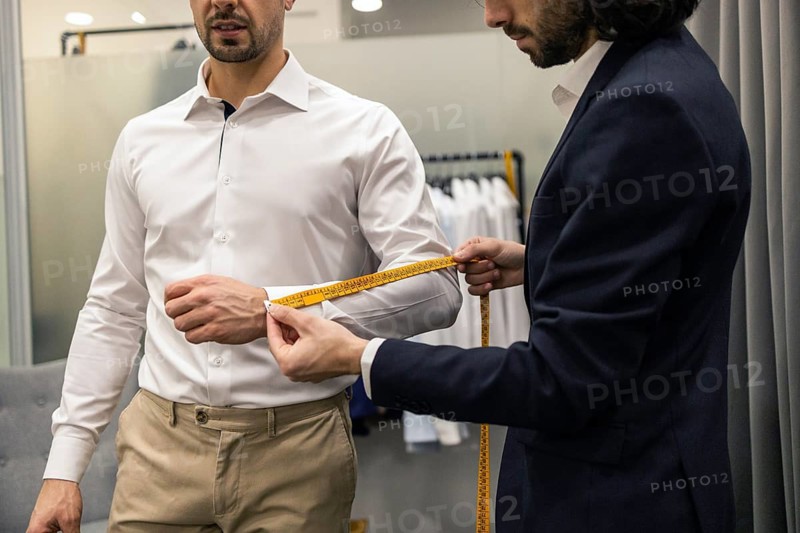 A tailor measures a client sleeve length with a yellow tape in a suit store. The client wears a white shirt and beige pants, representing precision and bespoke tailoring