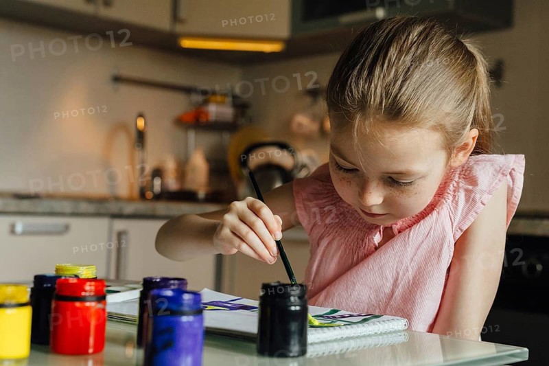 A young girl concentrates on painting with vibrant watercolors, surrounded by various paint jars on a kitchen table. The scene captures creativity and focus in a cozy home setting