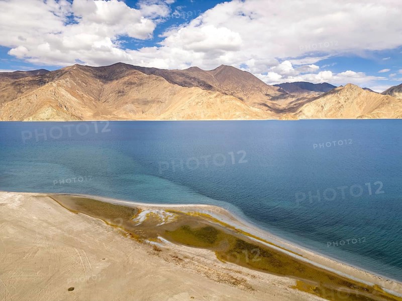 Pangong Tso lake in Ladakh, India, showcases its pristine blue waters embraced by rugged Himalayan mountain peaks, under a vivid sky of scattered clouds, creating a tranquil scene