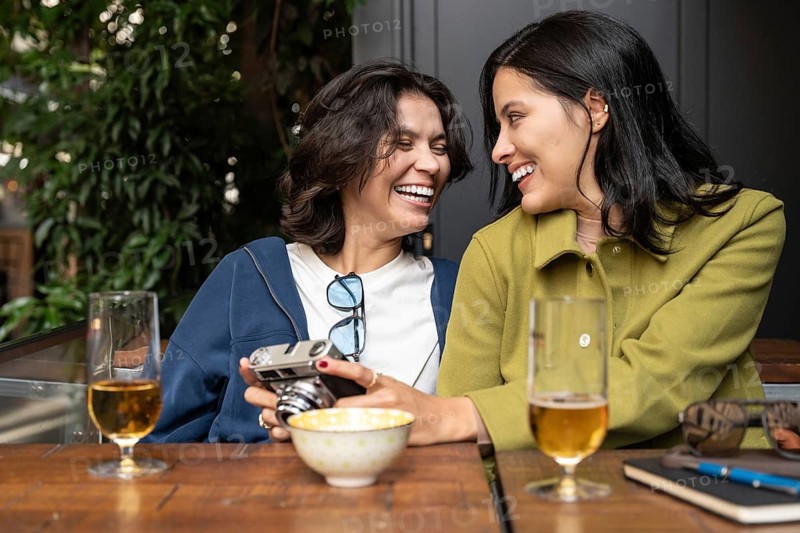 A cheerful lesbian couple sharing a moment of joy and laughter at an outdoor café. They are seated at a wooden table with drinks and a camera, enjoying each other's company