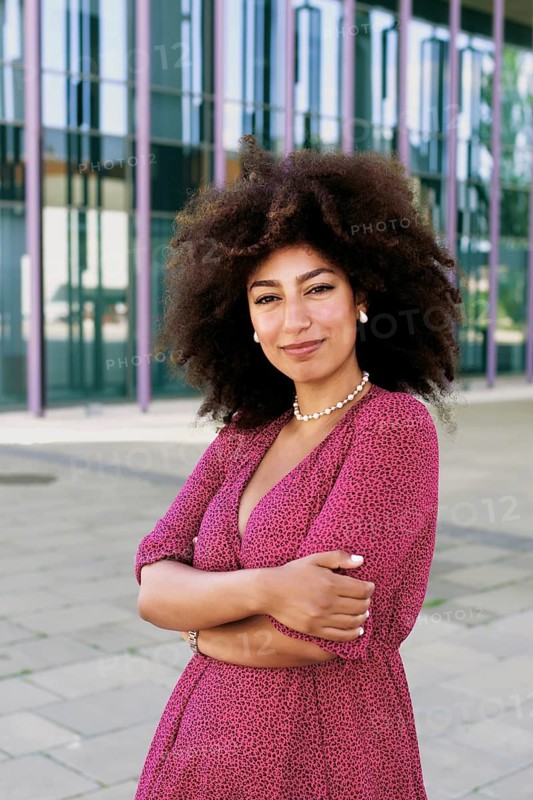 Young Arab woman with vibrant afro hair stands confidently looking at camera with crossed arms in front of a modern building. She wears a stylish pink dress, exuding elegance and self-assurance