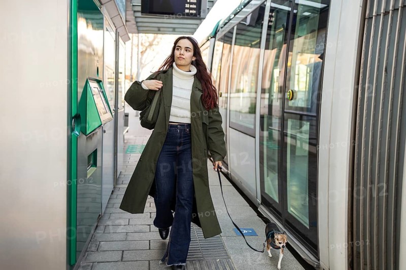 Woman walks her chihuahua at a tram stop, wearing a green coat and jeans. Urban setting with public transport, reflecting everyday city life and style