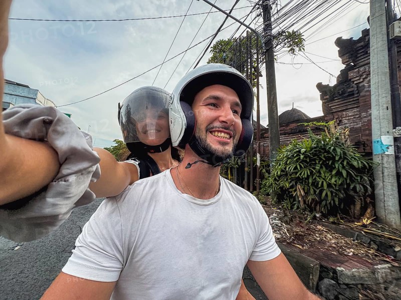 Smiling couple on a motorcycle ride, wearing helmets, capturing a happy moment on a selfie. Lush greenery and a clear sky create a perfect adventure backdrop