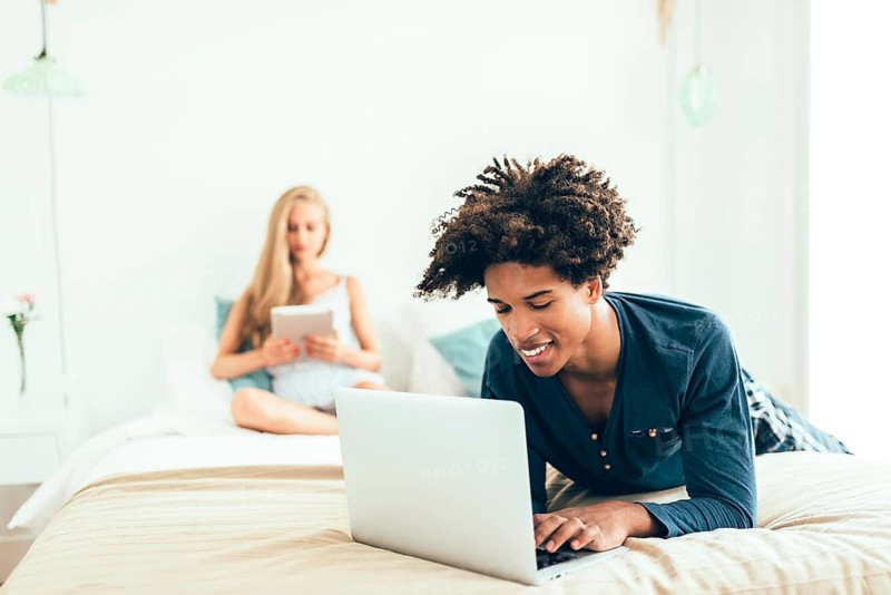 A young man lies on a bed using a laptop, while a woman sits in the background with a tablet. The scene captures a cozy, tech-driven ambiance in a bright bedroom