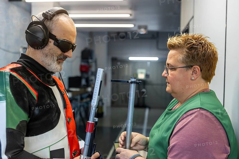 A blind athlete prepares for an Olympic shooting event with the help of a coach. Wearing protective gear and using specialized equipment, they focus on perfecting their technique