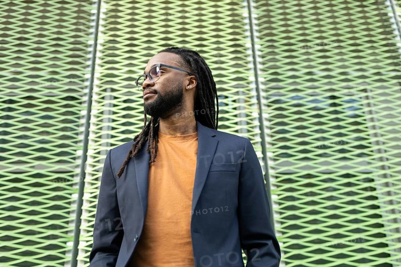 A cuban business man with long hair in glasses and a suit stands against a textured, patterned wall, embodying professionalism and style in an urban setting