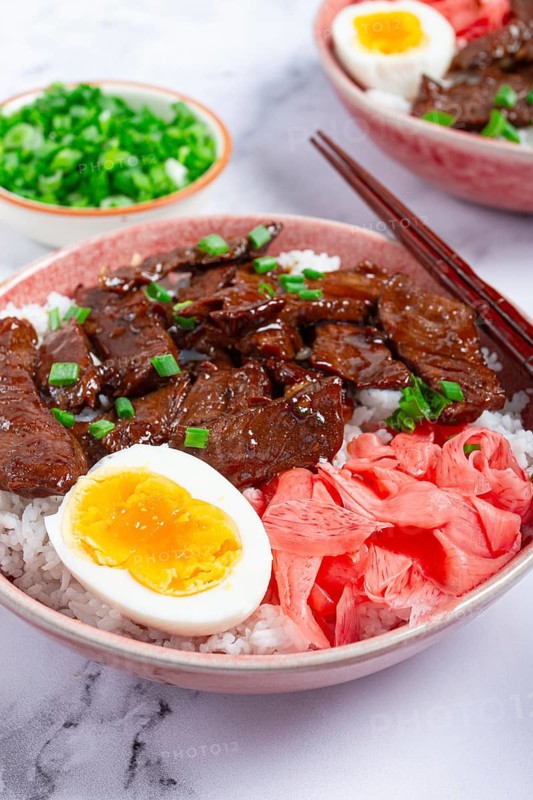 Gyudon Japanese beef with rice, pickled ginger, boiled egg slice, garnished with green onions, in a bowl with chopsticks