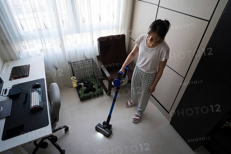 A woman efficiently vacuums the floor of a tidy home office, featuring a computer desk and a small pet area by the window, ensuring a clean and organized environment