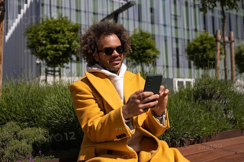 Afro man wearing sunglasses and a bright yellow coat sits on a bench, enjoying the outdoors and using his phone. A modern building is visible in the background