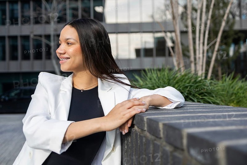 A smiling Hispanic woman in a white blazer enjoys a break outside a modern office building, radiating confidence and success in her business attire