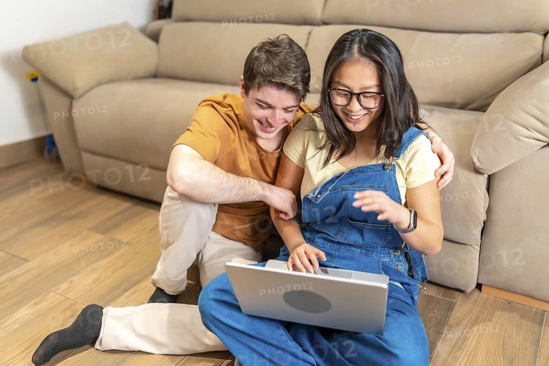 Happy multiethnic couple sitting on the floor and using laptop, surfing the web and having fun