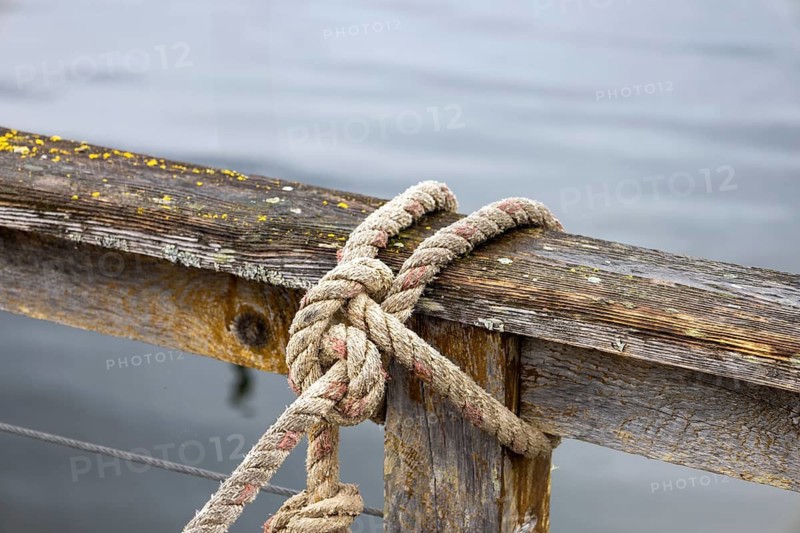 Detailed image of an old, weathered wooden dock with a sturdy rope tied in a knot, set against a backdrop of tranquil water in Hoonah, Alaska, a small town predominantly inhabited by the Tlingit community