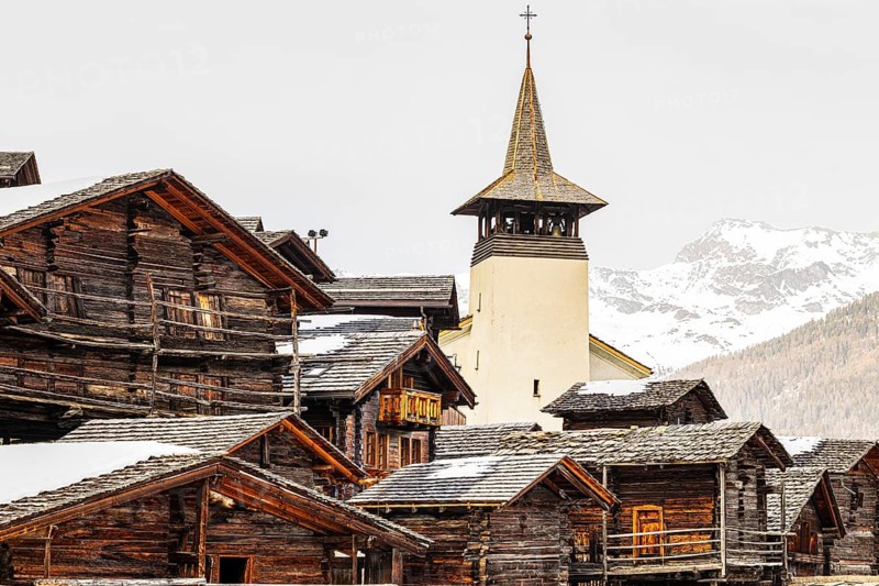 Snow-covered roofs of old farmhouses and barn storage, behind the church tower of the Saint Theodule church, Grimentz, Val d'Anniviers, Valais Alps, Canton of Valais, Switzerland