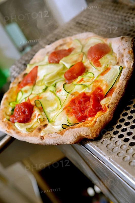 A freshly baked vegetable pizza topped with zucchini, tomato, and cheese rests on a metallic oven tray, steaming hot and ready to be served