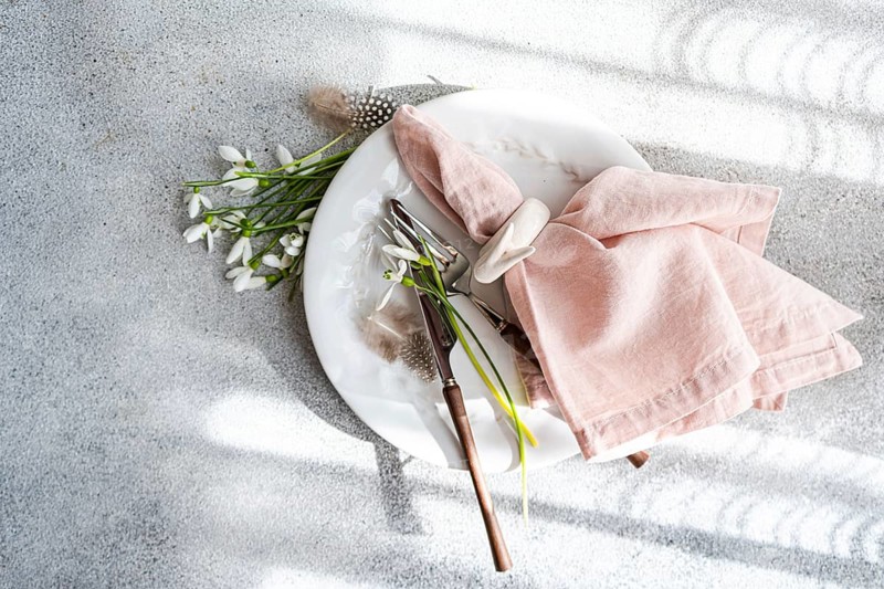From above, an elegant table setting featuring spring snowdrops, pink napkin, and vintage cutlery on a white plate, on a sunlit textured table