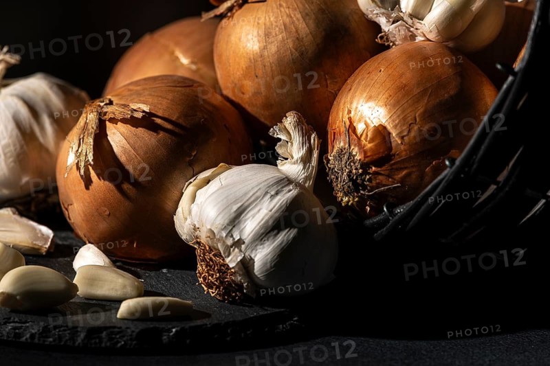 Onions and garlic next to a basket, dramatically lit on a dark background
