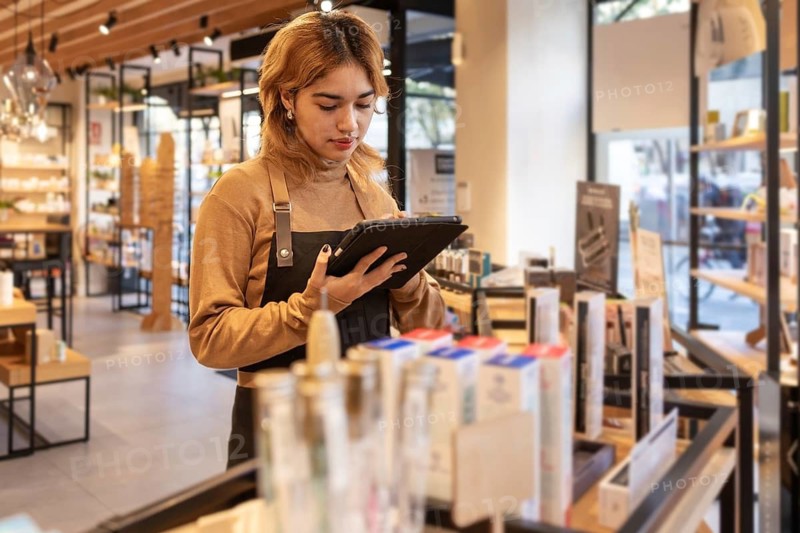 A cosmetics store worker in a brown sweater and black apron uses a tablet to check inventory. The store has well-organized shelves with various beauty products