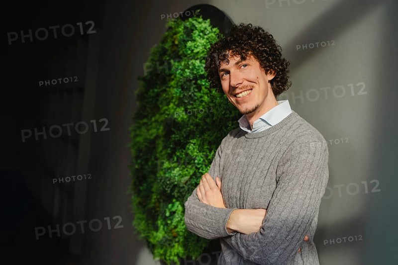 A young man with curly hair stands in front of a green wall, symbolizing themes of ecology and sustainable development. His casual attire and smile enhance the environmental theme