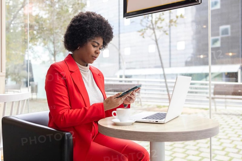 A woman in a red suit works on her phone and laptop in a modern cafe, reflecting a professional lifestyle. The setting is bright, emphasizing focus and efficiency