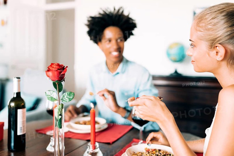 A romantic dinner scene with two people enjoying wine and a meal. A red rose and candle set the mood, emphasizing intimacy and warmth in the atmosphere