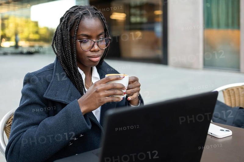An African American businesswoman with braids enjoys a coffee break outdoors while working remotely on her laptop