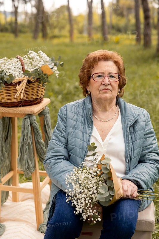 An elderly woman sits in an outdoor setting holding wildflowers with a serene expression. A wicker basket filled with flowers sits nearby, highlighting a peaceful moment in nature