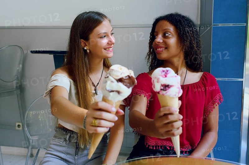 Two teenage girls, one Caucasian and the other African American, happily share ice cream cones during a friendly outing. They are having a casual conversation in a brightly lit setting