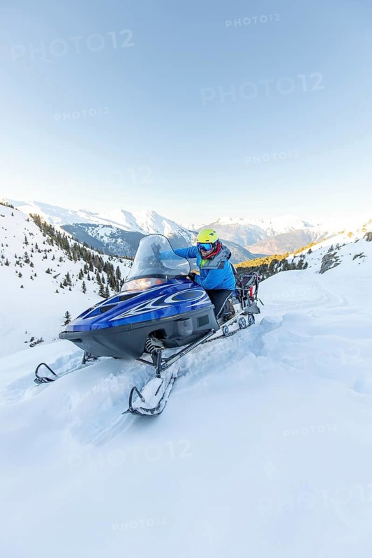 A thrilling snowmobile ride in a pristine winter wonderland. A man in a blue jacket and yellow helmet speeds through a snowy mountain landscape under a clear blue sky