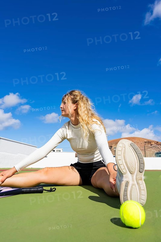 Woman with blond hair, in a post-game stretching routine on a tennis court. She sits comfortably, extending her leg while gently leaning forward, away from the camera, under a clear, sunny sky