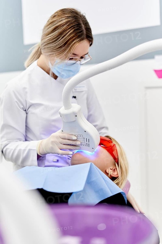 A dentist performs a teeth whitening procedure in a modern dental clinic. The patient wears protective glasses while a special device emits blue light on their teeth