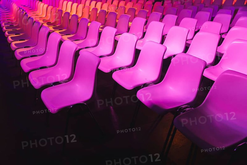 A captivating array of pink chairs in neat rows, lit by vivid magenta lighting at a conference of AI in Amsterdam. The dark background enhances the vibrant and stylish atmosphere