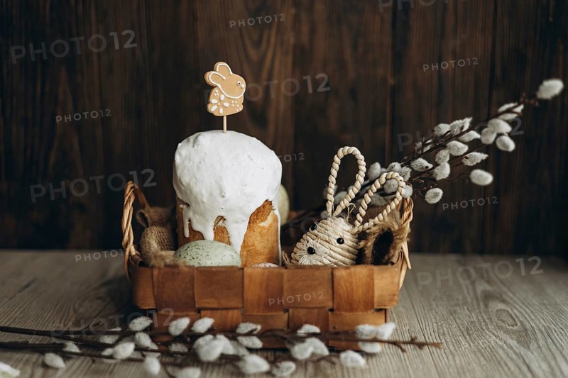 An ornate setup displaying traditional Russian Easter bread, Kulich, garnished with a gingerbread cookie bunny and surrounded by basket and willow catkin branches
