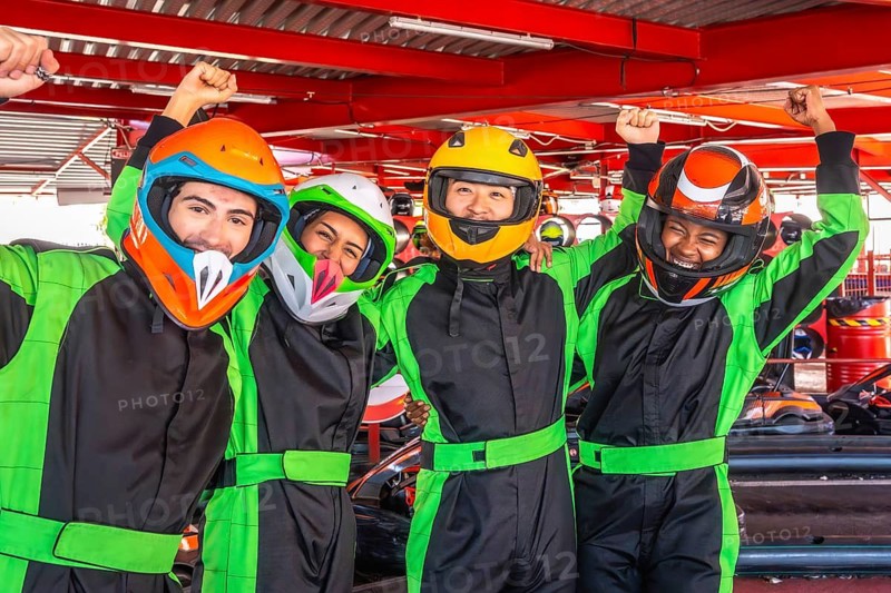 Group of diverse young adults wearing colorful helmets and racing suits, smiling and raising arms in celebration after an exciting karting race at an indoor track
