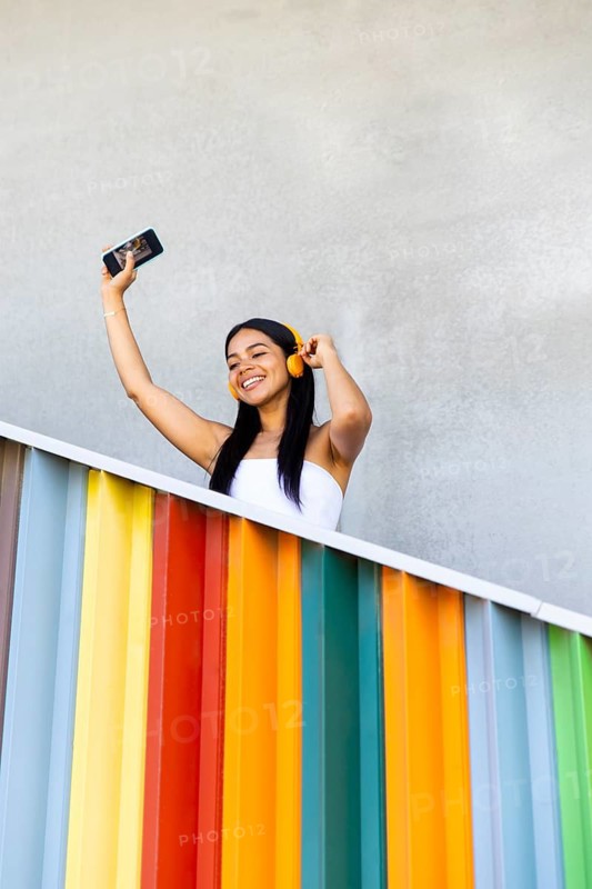 A woman wearing vibrant headphones enjoys music while standing against a backdrop of colorful modern architecture. She holds a smartphone, embracing a moment of joy outdoors