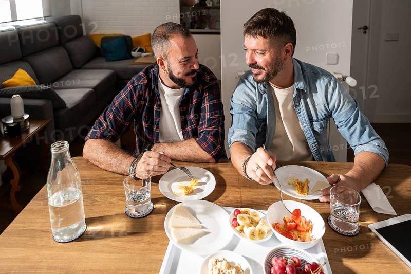 A gay couple enjoying breakfast together at home, sharing a moment of laughter and connection. Sunlight gently illuminates the table set with a healthy meal