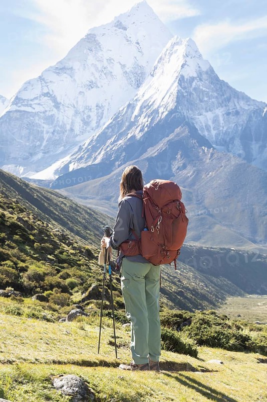 A woman in hiking gear admires the stunning view of the Himalayas, including Mount Everest, during a trek. She carries a large backpack and uses trekking poles