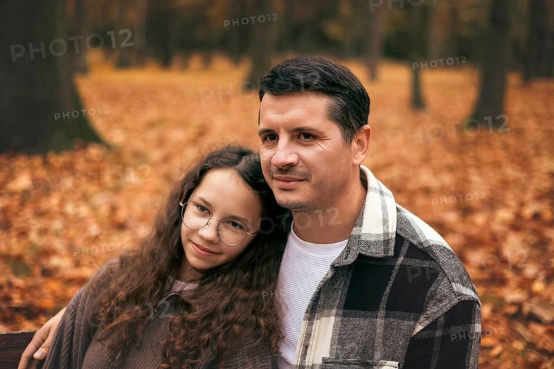 A father and daughter stand together in an autumn park, surrounded by vibrant fallen leaves. The warm atmosphere highlights their bond and the beauty of the season
