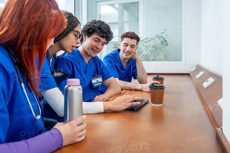 Diverse group of medical and nursing students in scrubs and stethoscopes collaborating over a tablet during a clinic break, focused on teamwork, learning and clinical training