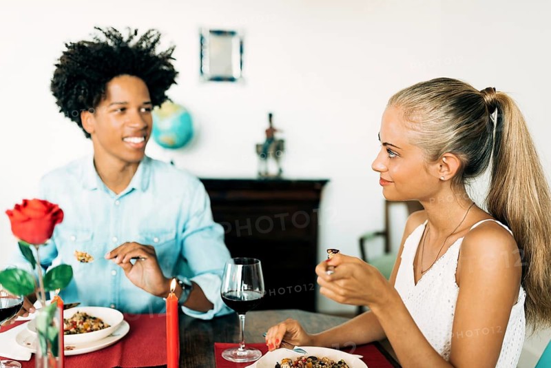 A young couple shares a romantic dinner, surrounded by red wine, candles, and a single rose, creating a warm and intimate atmosphere for a cozy evening together