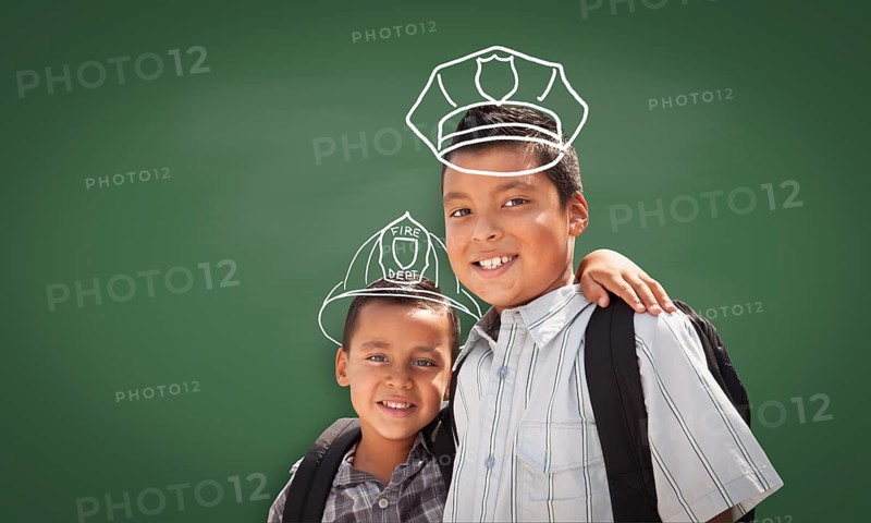 Young Hispanic Student Boy Wearing Backpack Front Of Blackboard with Fireman Helmet And Policeman Hat Drawn In Chalk Over Heads