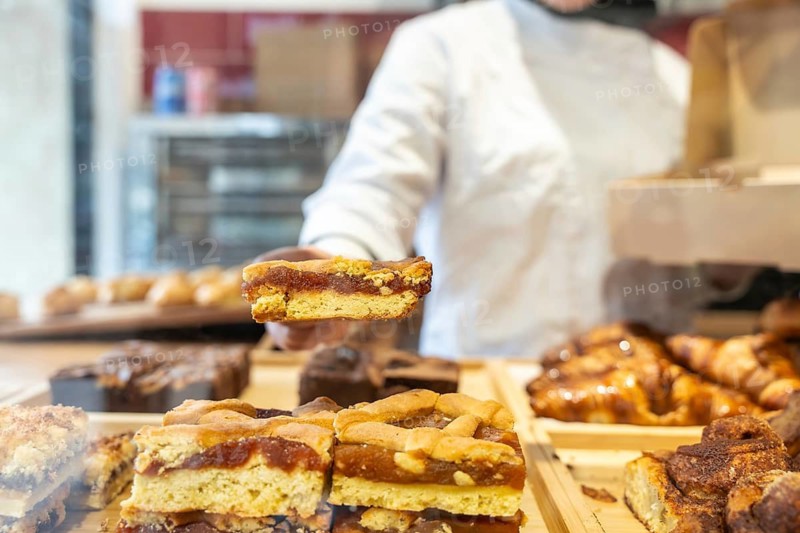Baker in white uniform offering a slice of fruit tart with jam filling to a customer at a bakery storefront, pastries displayed on the counter, fresh and handmade ambiance